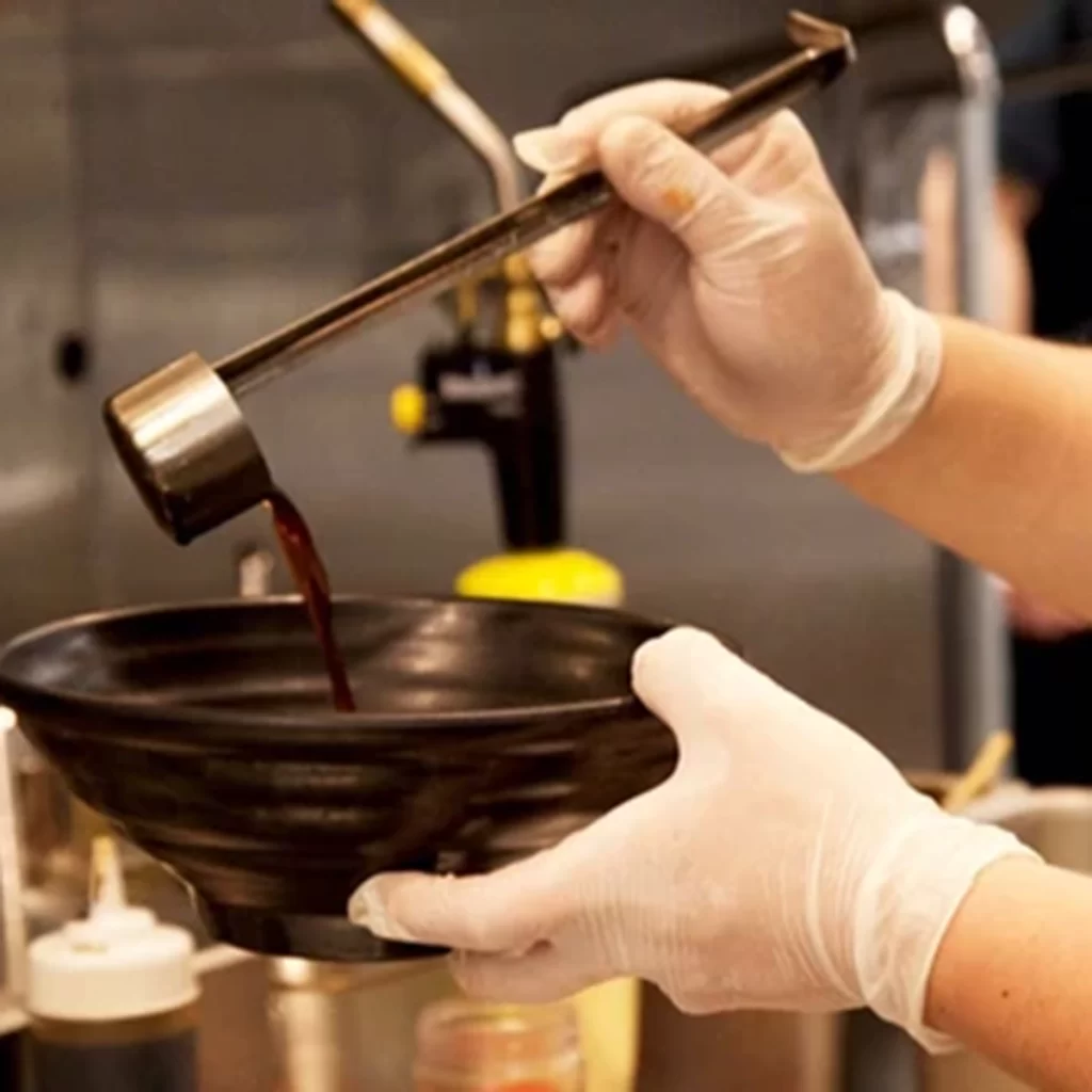 Chef pours dark seasoning sauce into ramen bowl at Ichiddo Ramen, Ramen Restaurant in Poughkeepsie